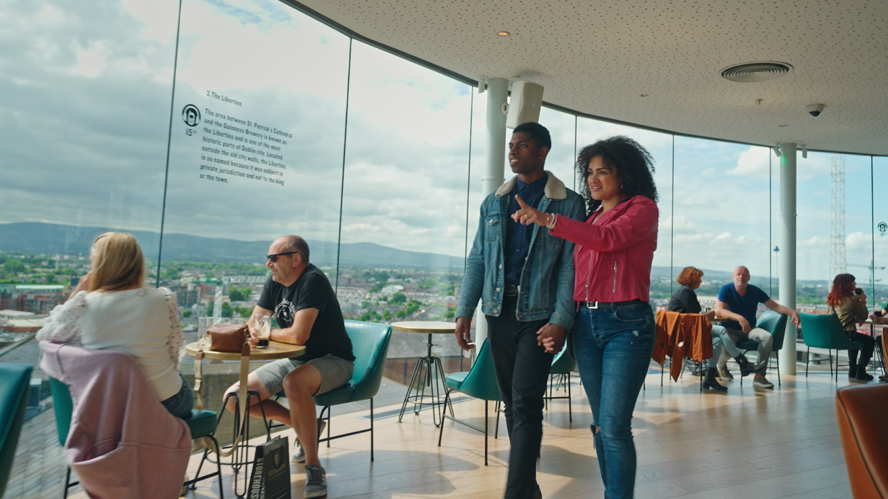 a man and a woman in the guinness bar walking through, other people sitting down at tables 